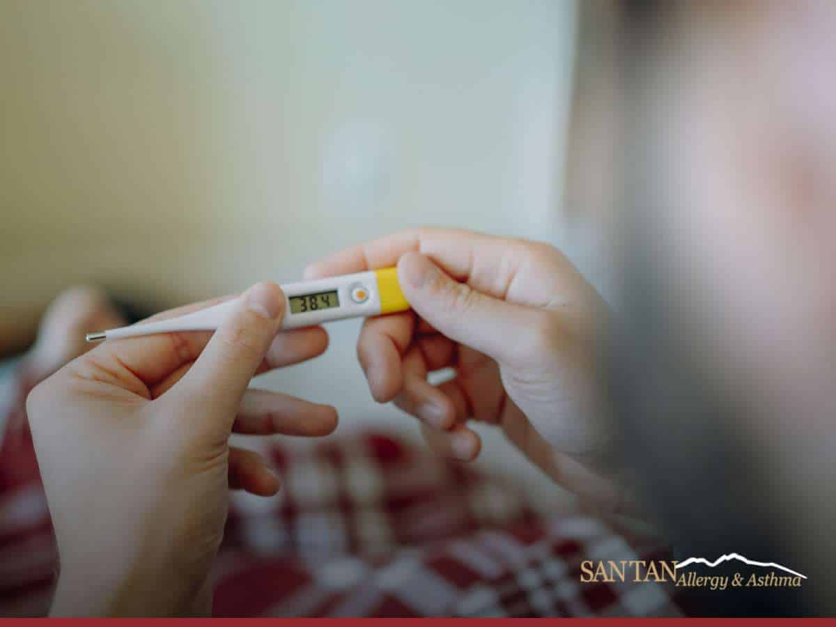 Digital thermometer reading 38.4°C held by a person, with the San Tan Allergy & Asthma logo, symbolizing consultation with allergic experts.